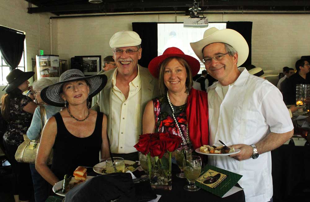 Group of two older men and two older women standing at a small table with plates of food, all wearing different types of hats.