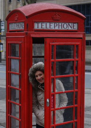 Isla Waite in a light-colored parka, standing inside and poking her head out from behind the door of a red telephone booth in England.