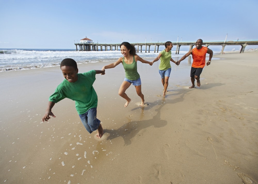 Three young adults and one older child with hands linked and running along the sandy shore at the beach on a sunny day. There is a pier in the distance.