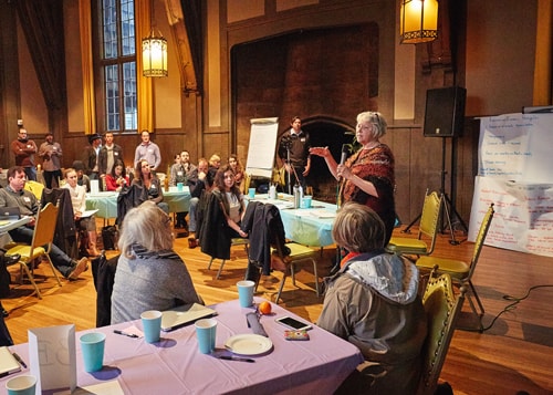 An older woman with short gray hair stands and addresses a mixed group of adults sitting at tables.