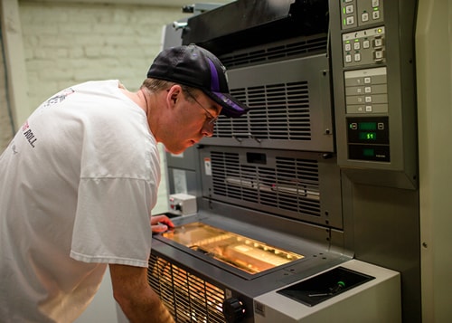 A man in glasses, a white t-shirt and a dark cap operates large printing machinery