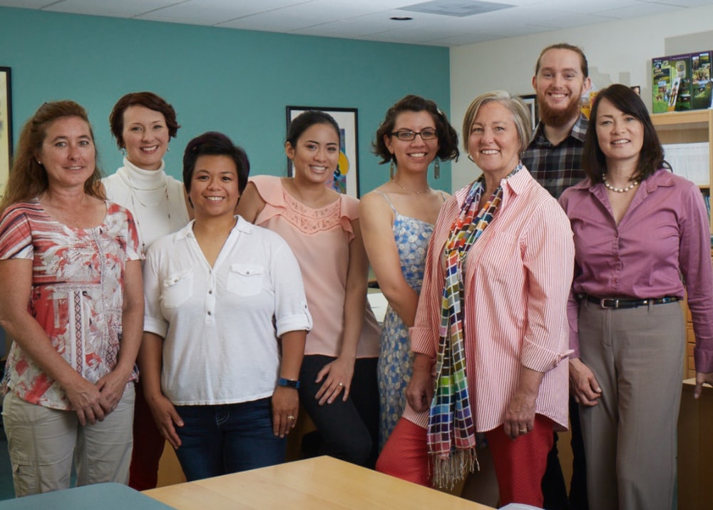 Group of seven adult women and one adult man standing close together in an office setting and smiling at the camera.