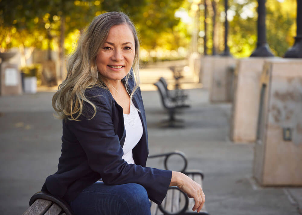 A middle-aged woman with long wavy hair wearing a white top, dark blue blazer, and jeans, sitting on a park bench while leaning forward with her elbow resting on her knee.