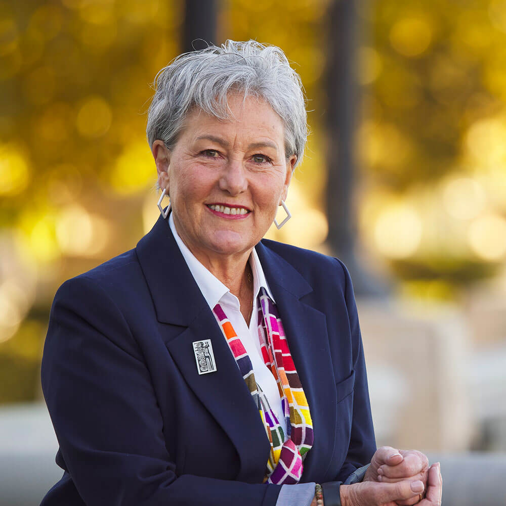 An older woman with short silver hair wearing a white collared shirt, dark blue blazer, and a multi-colored scarf with a square pattern.