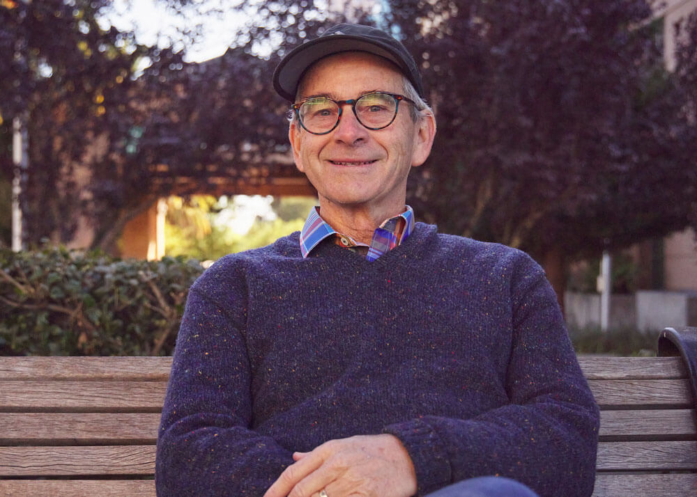And older man sitting on a bench, wearing glasses, a dark cap, and a dark blue sweater.