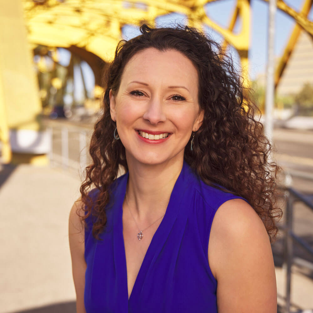 A woman with long curly dark hair and dark eyes wearing a short-sleeved indigo top, with a yellow bridge in the background.