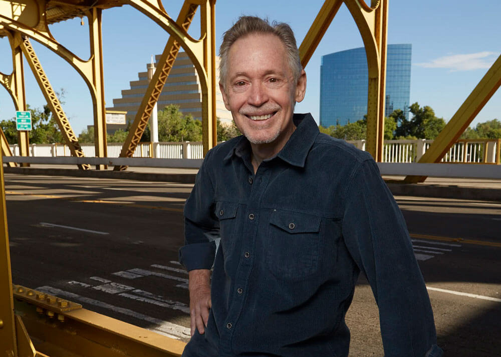 An older man with gray hair and a moustach wearing a dark button-up shirt and leaning against a yellow bridge railing.