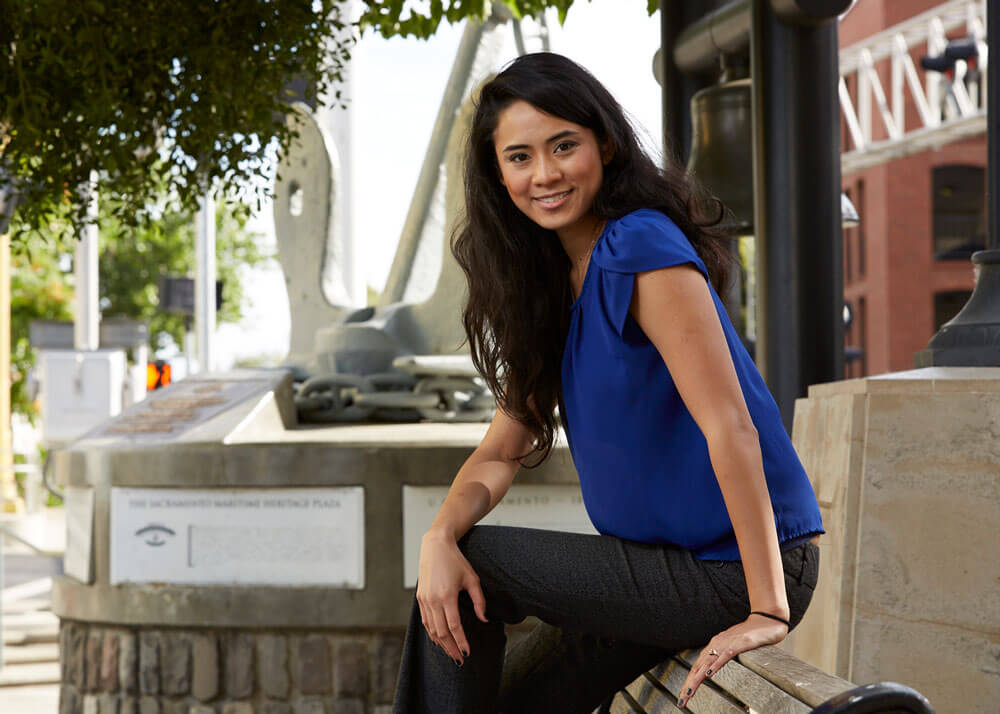 A young woman with light brown skin and long dark hair, wearing a blue top and dark pants, sitting on top of the back of a park bench.
