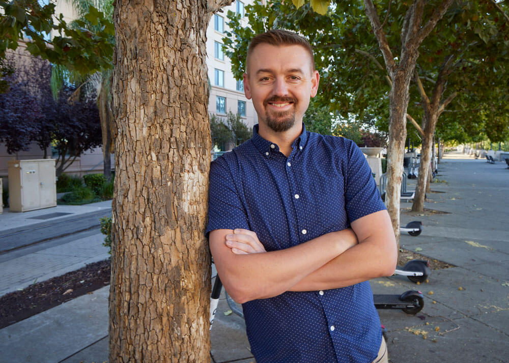 A man in his thirties with short brown hair, a short bead and moustache, wearing a button-up blue shirt with his arms folded and leaning against a small tree trunk.